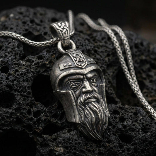 Oxidized Odin pendant necklace on textured volcanic rock, centered close-up highlighting the one-eyed helmeted face, braided beard, and deep antique shadowed contrast.