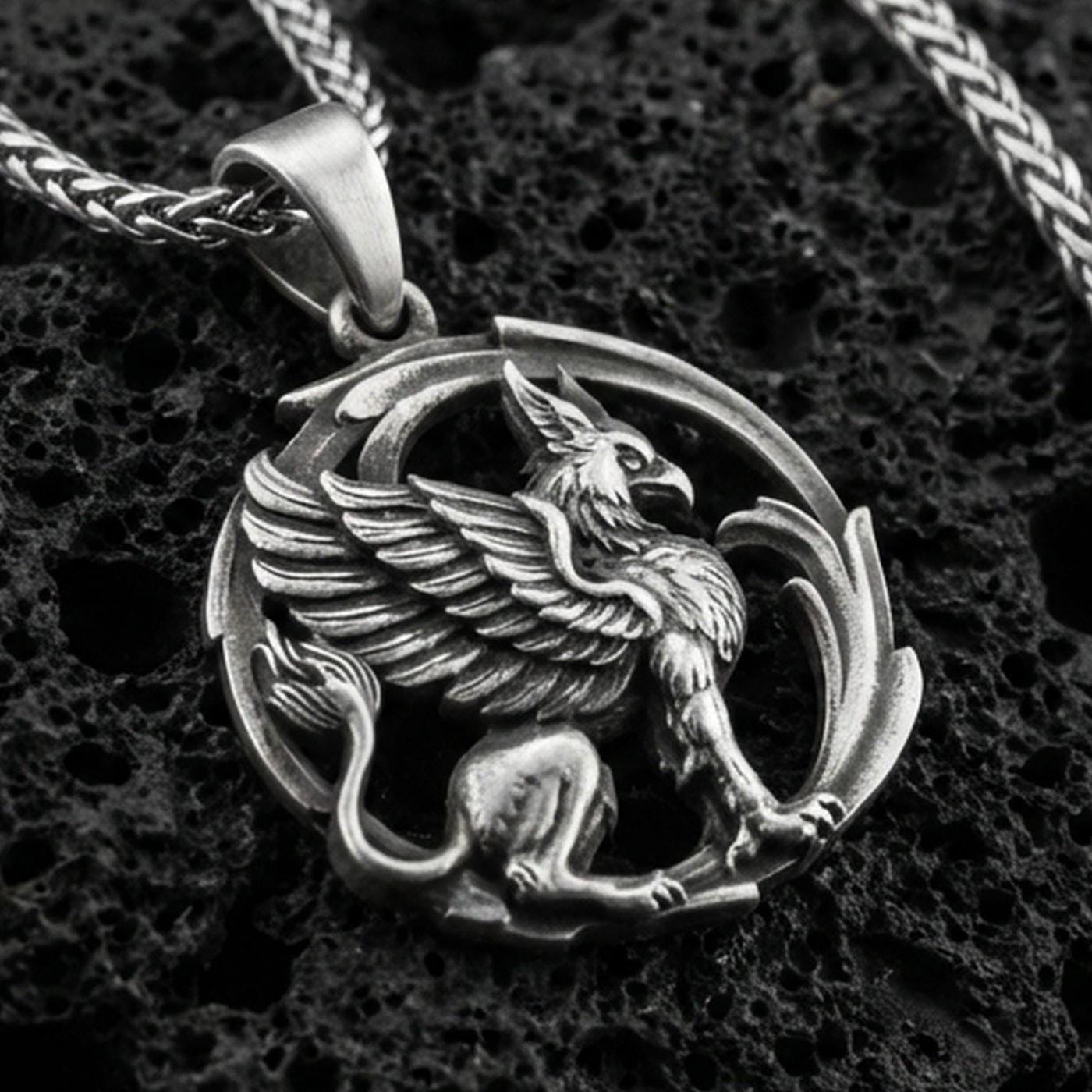 Oxidized griffin pendant necklace on textured volcanic rock, centered close-up highlighting the winged gryphon relief, curled tail, and deep antique shadowed contrast.