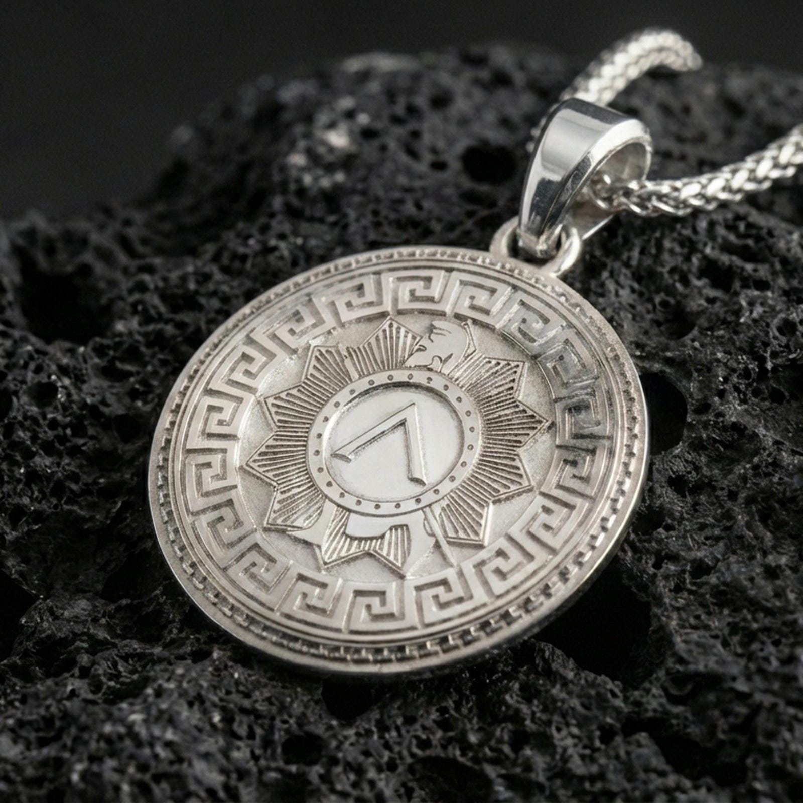 Polished silver Spartan shield medallion pendant necklace on textured volcanic rock, crisp macro product photo emphasizing clean edges, sharp geometric engraving, and bright reflective highlights.