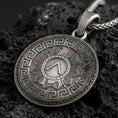 Load image into Gallery viewer, Oxidized Spartan shield medallion pendant necklace on textured volcanic rock, centered close-up highlighting the Greek key border, sunburst shield design, and deep antique contrast.
