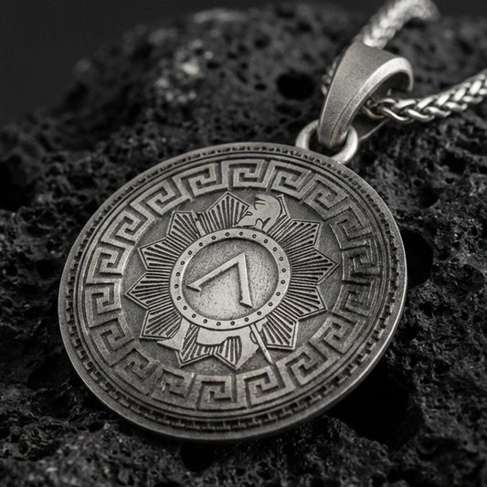 Oxidized Spartan shield medallion pendant necklace on textured volcanic rock, centered close-up highlighting the Greek key border, sunburst shield design, and deep antique contrast.