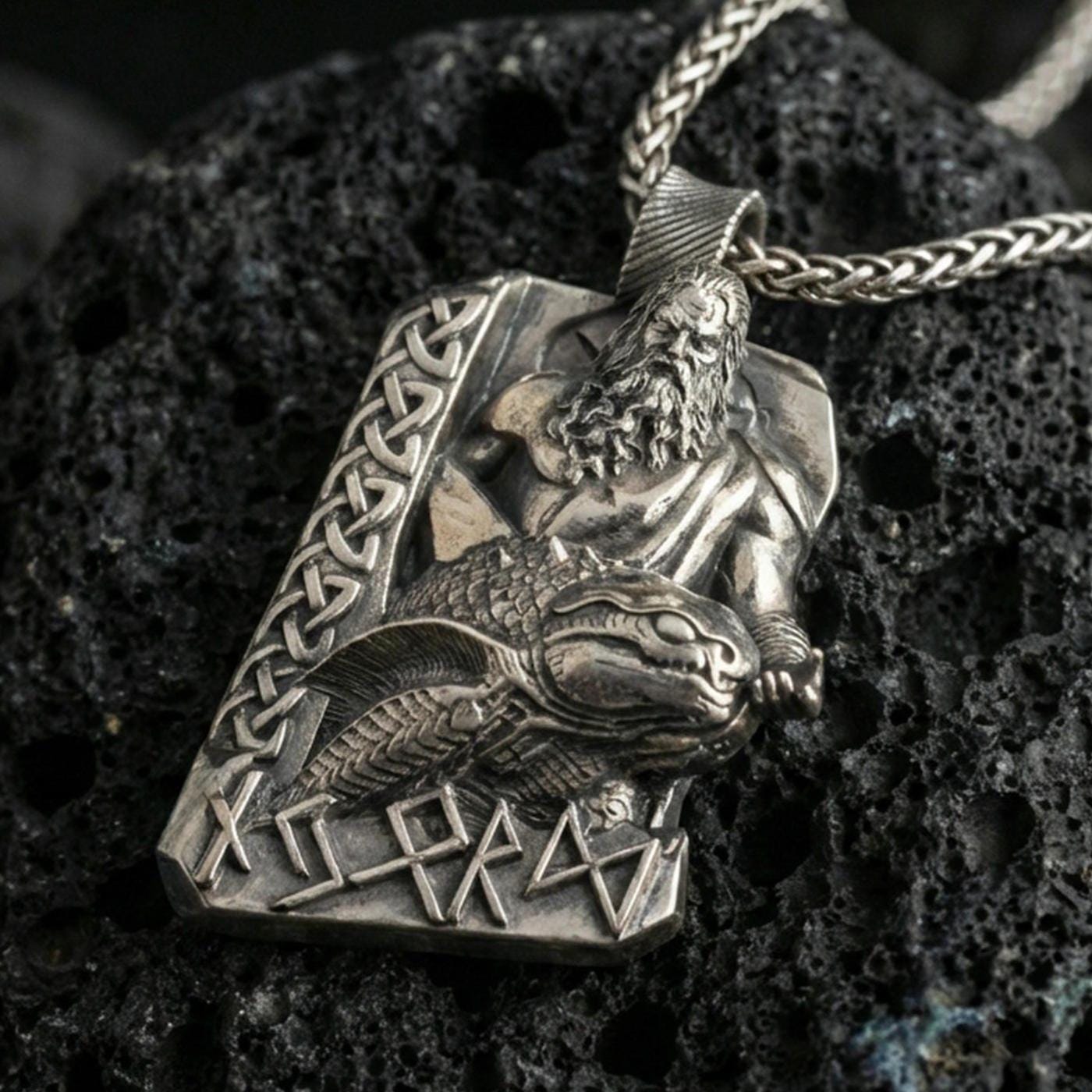 Oxidized Njord Norse sea god pendant necklace on textured volcanic rock, centered close-up highlighting the bearded deity relief, sea-creature motif, rune lettering, and deep antique contrast.
