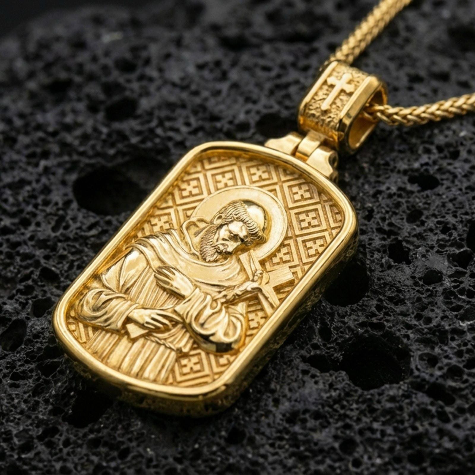 Gold plated Saint Francis pendant with sculpted saint portrait, cross motif, and engraved patterned background showcased on black volcanic rock in a refined macro jewelry image