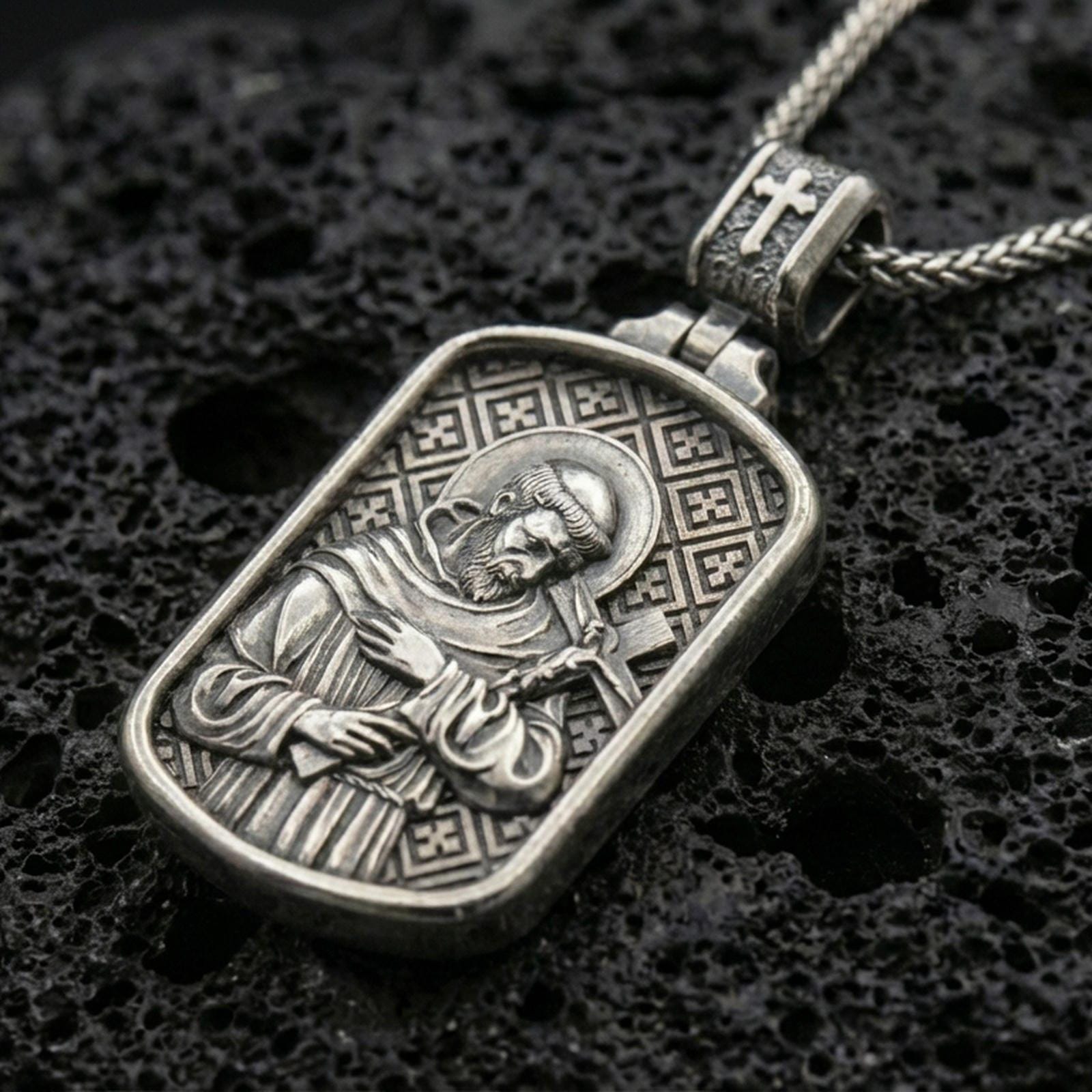 Oxidized Saint Francis pendant with halo, cross detail, and framed devotional portrait displayed on dark volcanic rock in a close-up luxury jewelry composition