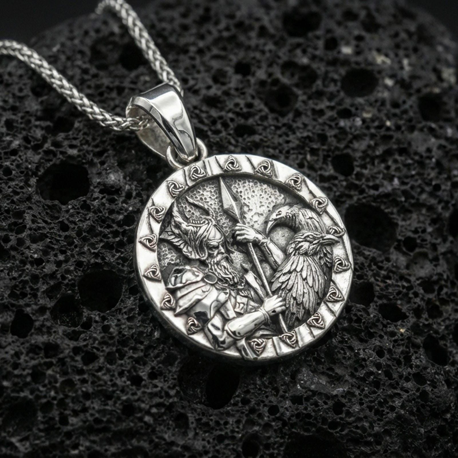 Polished silver Odin and raven medallion pendant necklace on textured volcanic rock, crisp macro product photo emphasizing clean edges, sharp feather engraving, and detailed Norse scene relief.