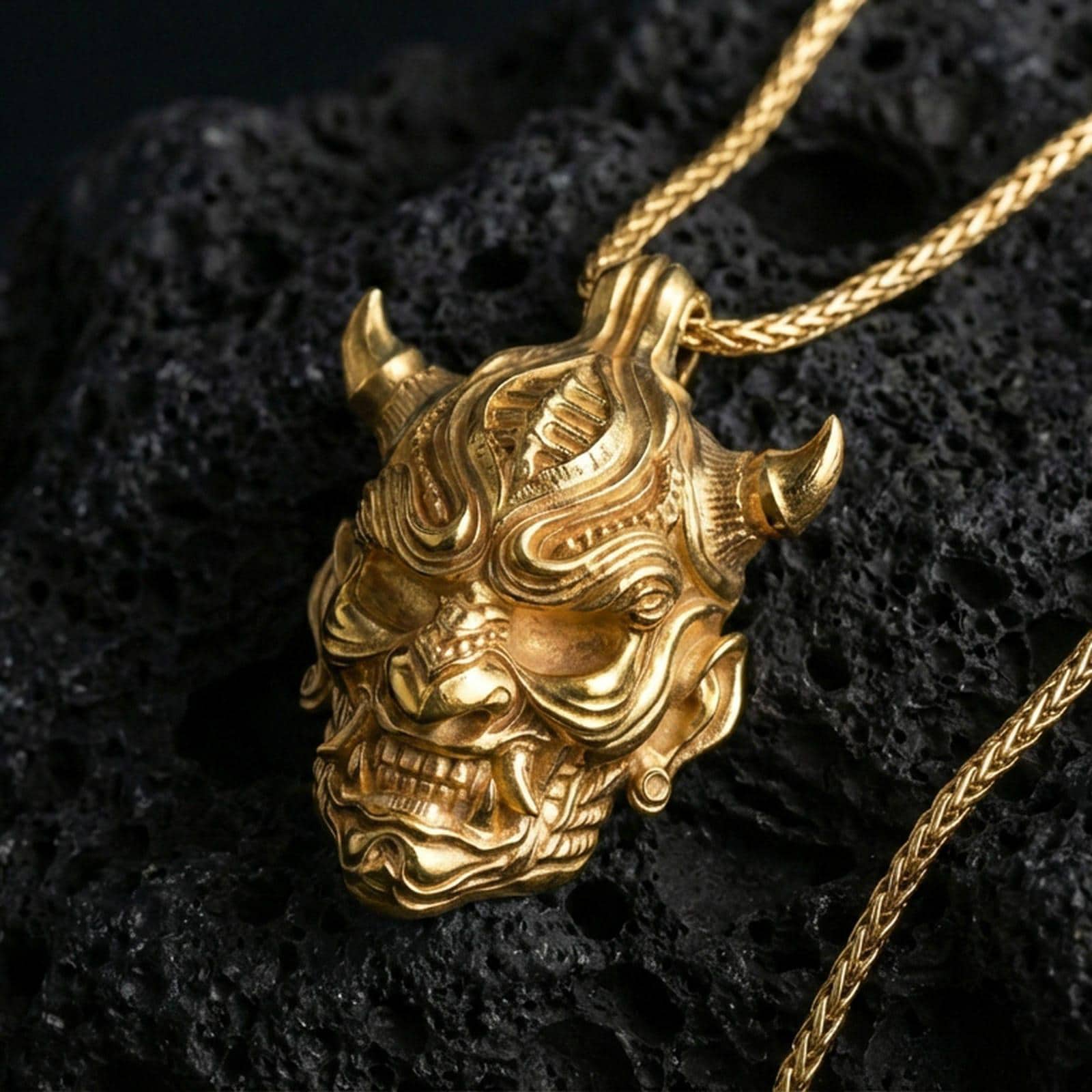 Gold plated oni mask pendant necklace on textured volcanic rock, premium close-up with warm reflective shine across the horns, fierce expression, and high-relief mask contours.