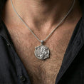 Load image into Gallery viewer, Polished silver Saint George medallion with horse and dragon relief worn on the neck in a close lifestyle frame, highlighting the detailed battle scene and Christian symbolism
