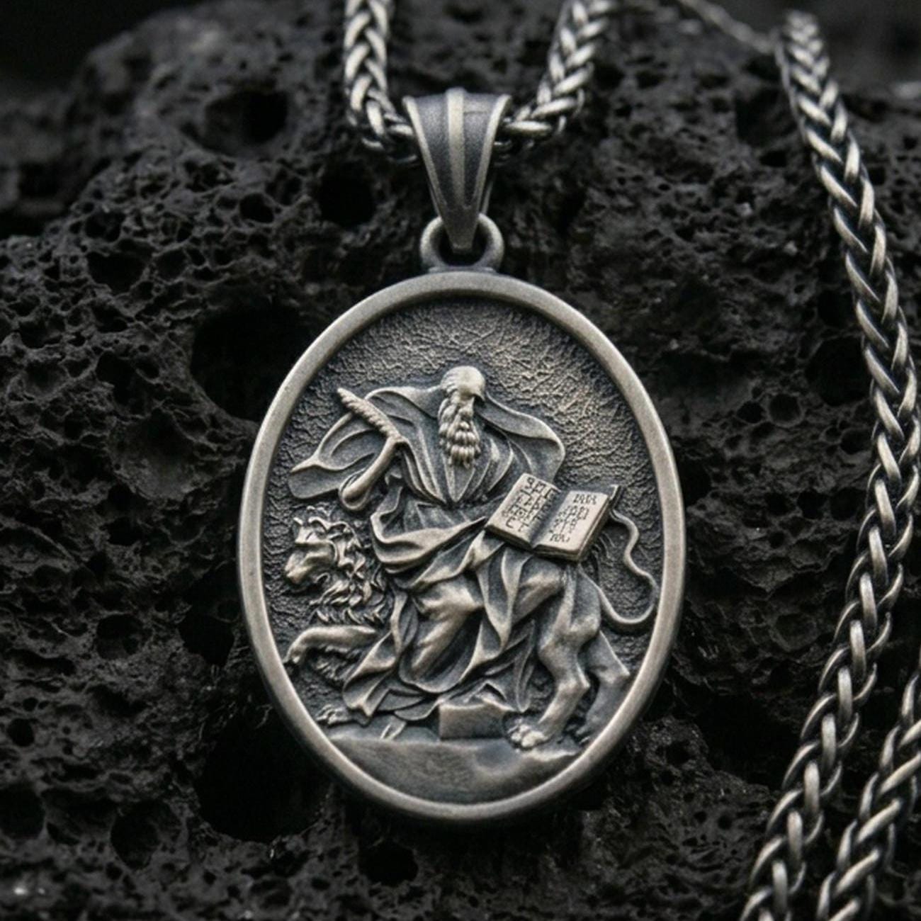 Oxidized saint medallion pendant with a seated bearded figure, lion detail, and open scripture relief displayed on dark volcanic rock in a close-up luxury jewelry composition