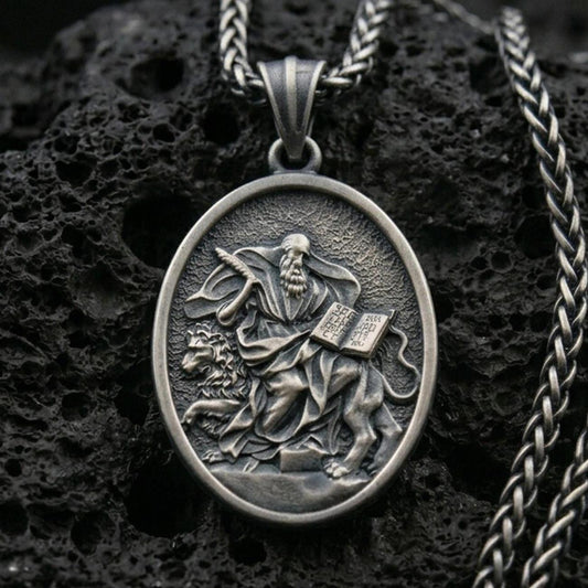 Oxidized saint medallion pendant with a seated bearded figure, lion detail, and open scripture relief displayed on dark volcanic rock in a close-up luxury jewelry composition