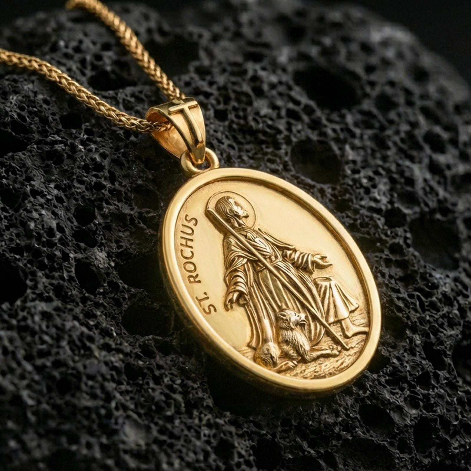 Gold plated Saint Rochus medal with detailed saint portrait, walking staff, and loyal dog motif showcased on black volcanic rock in a refined macro jewelry image