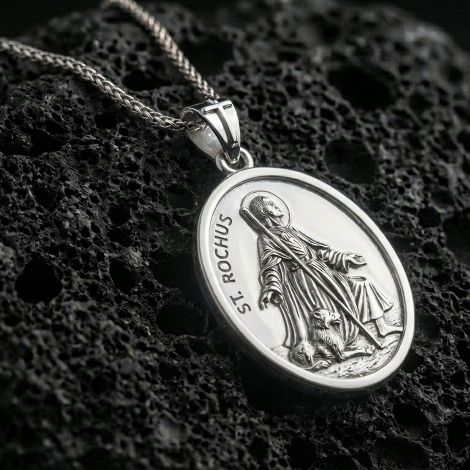 Polished silver Saint Rochus pendant with oval frame, devotional figure, and dog relief placed on textured volcanic rock in a premium studio product shot