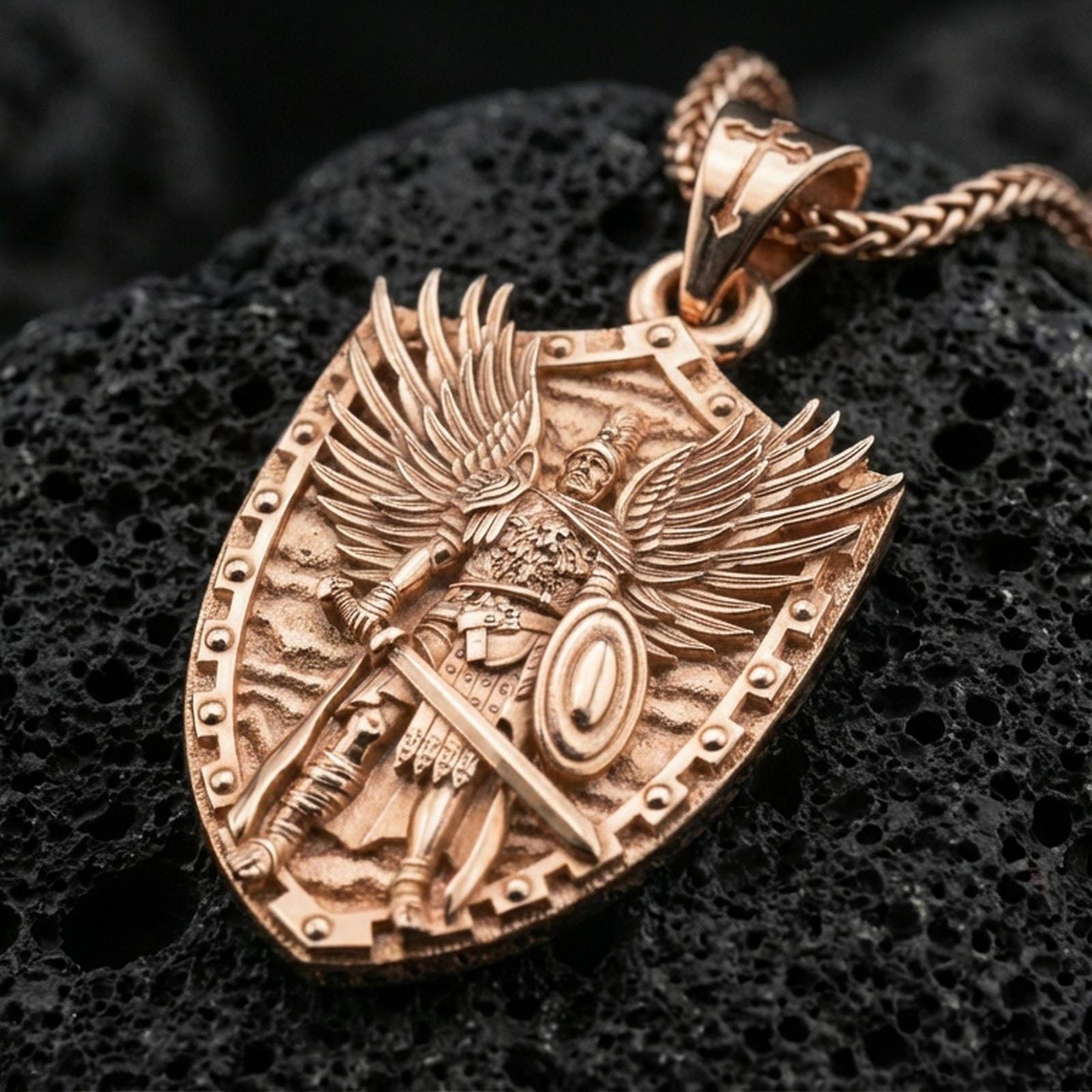Rose gold plated Saint Michael guardian pendant with winged warrior motif, shield border, sword, and armor relief resting on volcanic rock in an elegant close-up product presentation