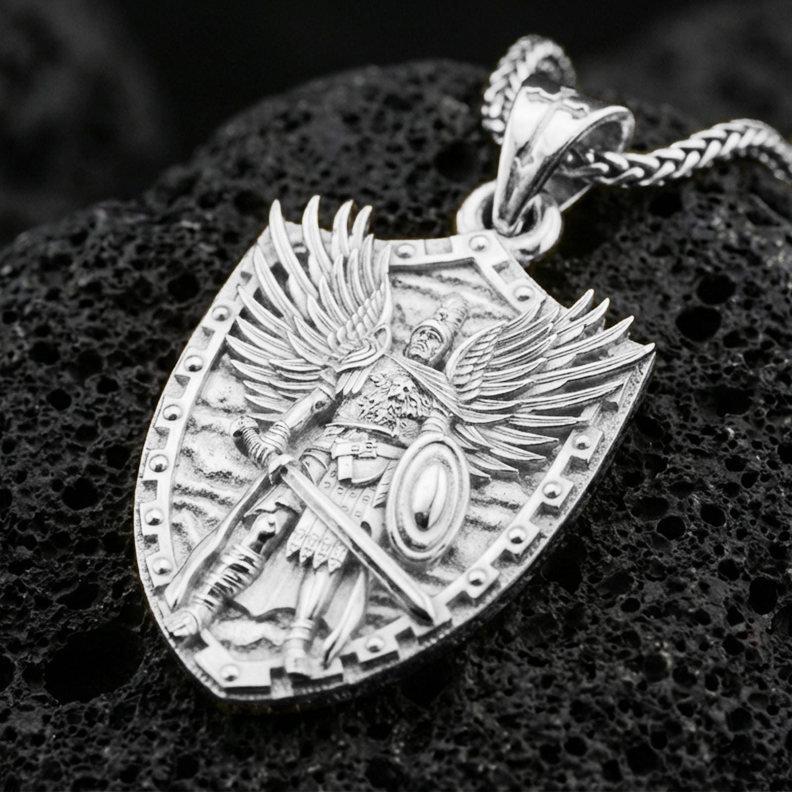 Polished silver Saint Michael pendant with angel warrior relief, shield frame, sword, and raised wing details placed on textured volcanic rock in a premium studio product shot