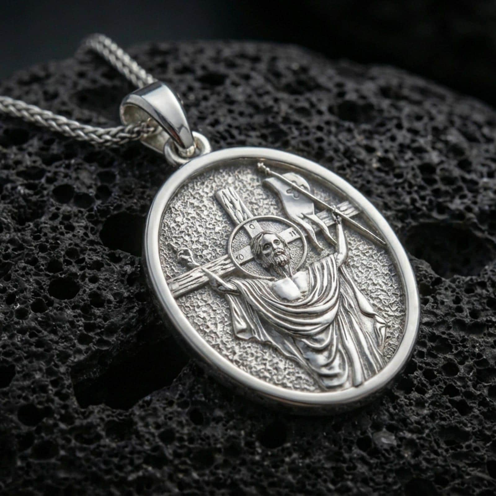 Polished silver religious oval pendant with resurrected Jesus figure, victory banner, and cross motif placed on textured volcanic rock in a premium studio product shot