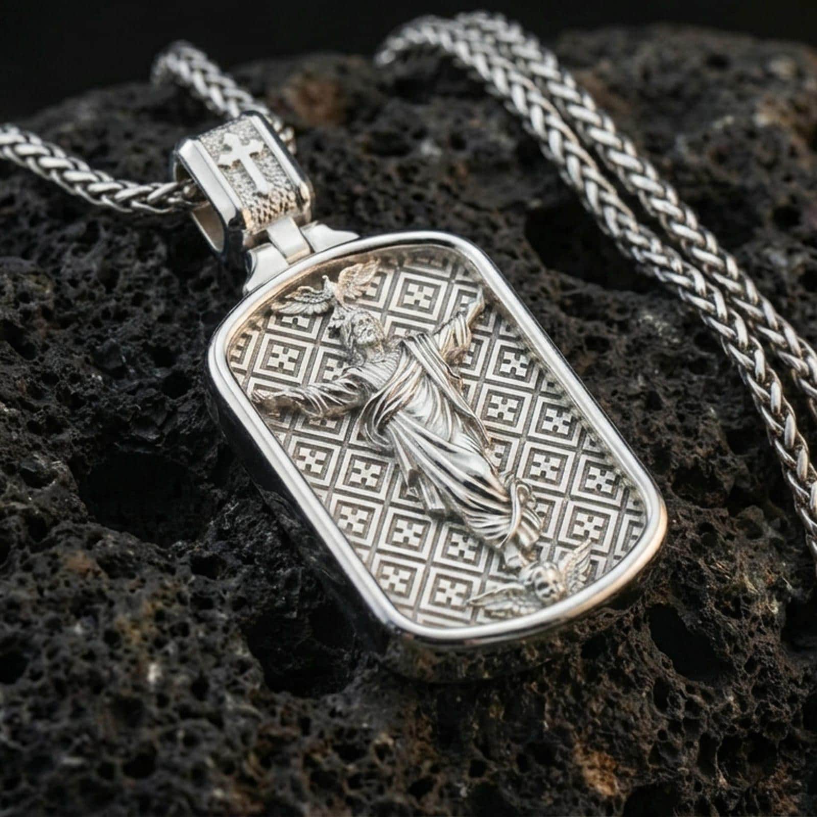 Polished silver Christian pendant with risen Jesus relief, geometric engraved background, and rounded rectangular frame placed on textured volcanic rock in a premium studio product shot