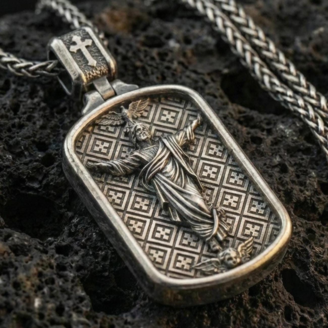 Oxidized Jesus pendant with ascending Christ figure, angel motifs, and framed devotional design displayed on dark volcanic rock in a close-up luxury jewelry composition