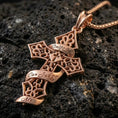 Load image into Gallery viewer, Rose gold plated ornate cross pendant with filigree cutout details, engraved ribbon lettering, and gothic silhouette resting on volcanic rock in an elegant close-up product presentation
