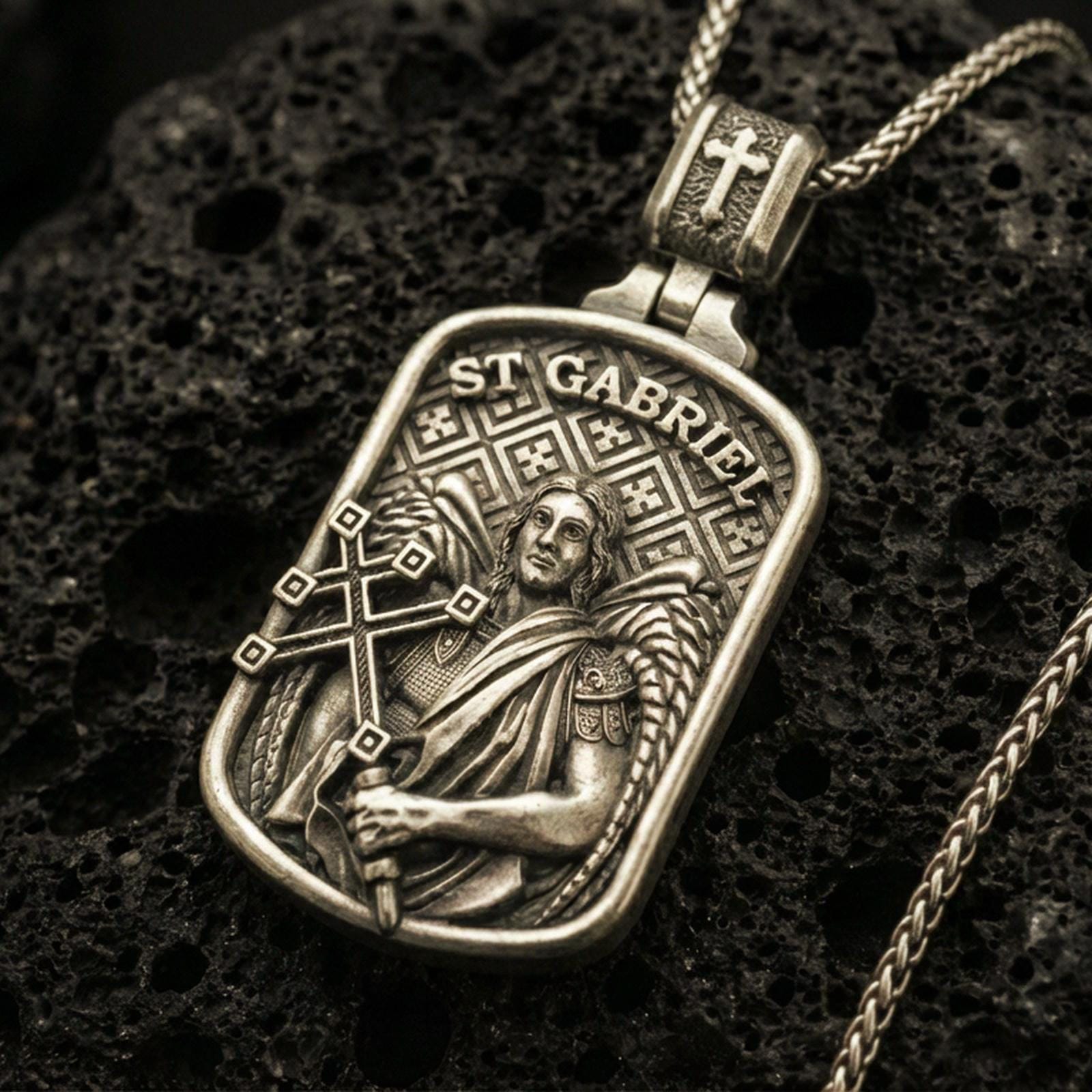 Bronze Saint Gabriel pendant with angelic figure, wing detail, and ST Gabriel inscription displayed on dark volcanic rock in a close-up luxury jewelry composition