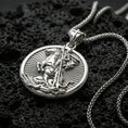 Load image into Gallery viewer, Polished silver Saint George medallion with horseman figure, circular frame, and radiant engraved background placed on textured volcanic rock in a premium studio product shot
