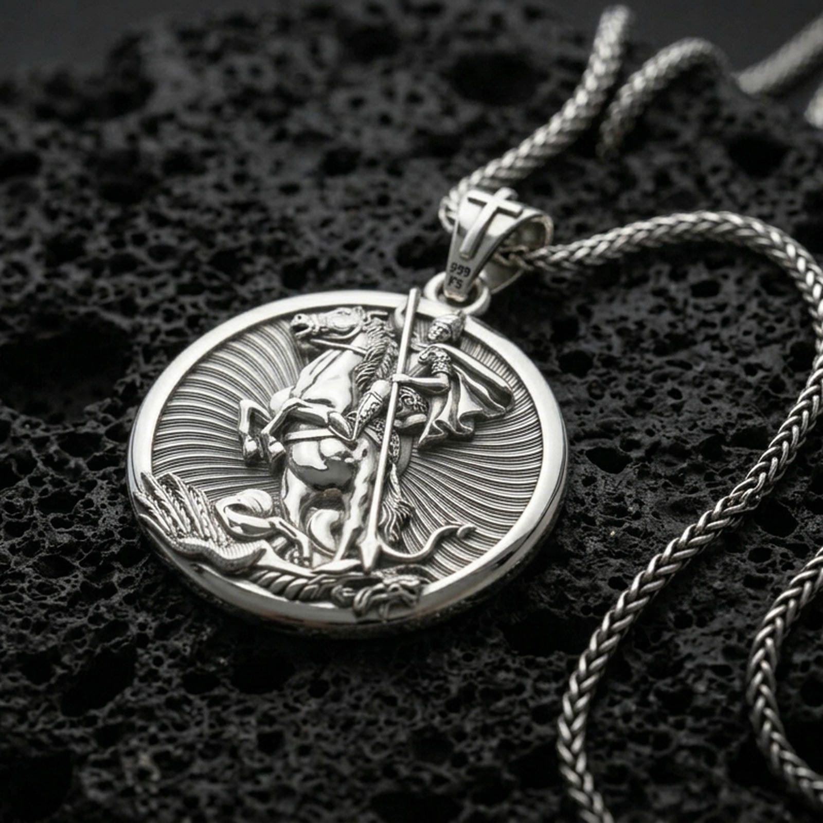Polished silver Saint George medallion with horseman figure, circular frame, and radiant engraved background placed on textured volcanic rock in a premium studio product shot