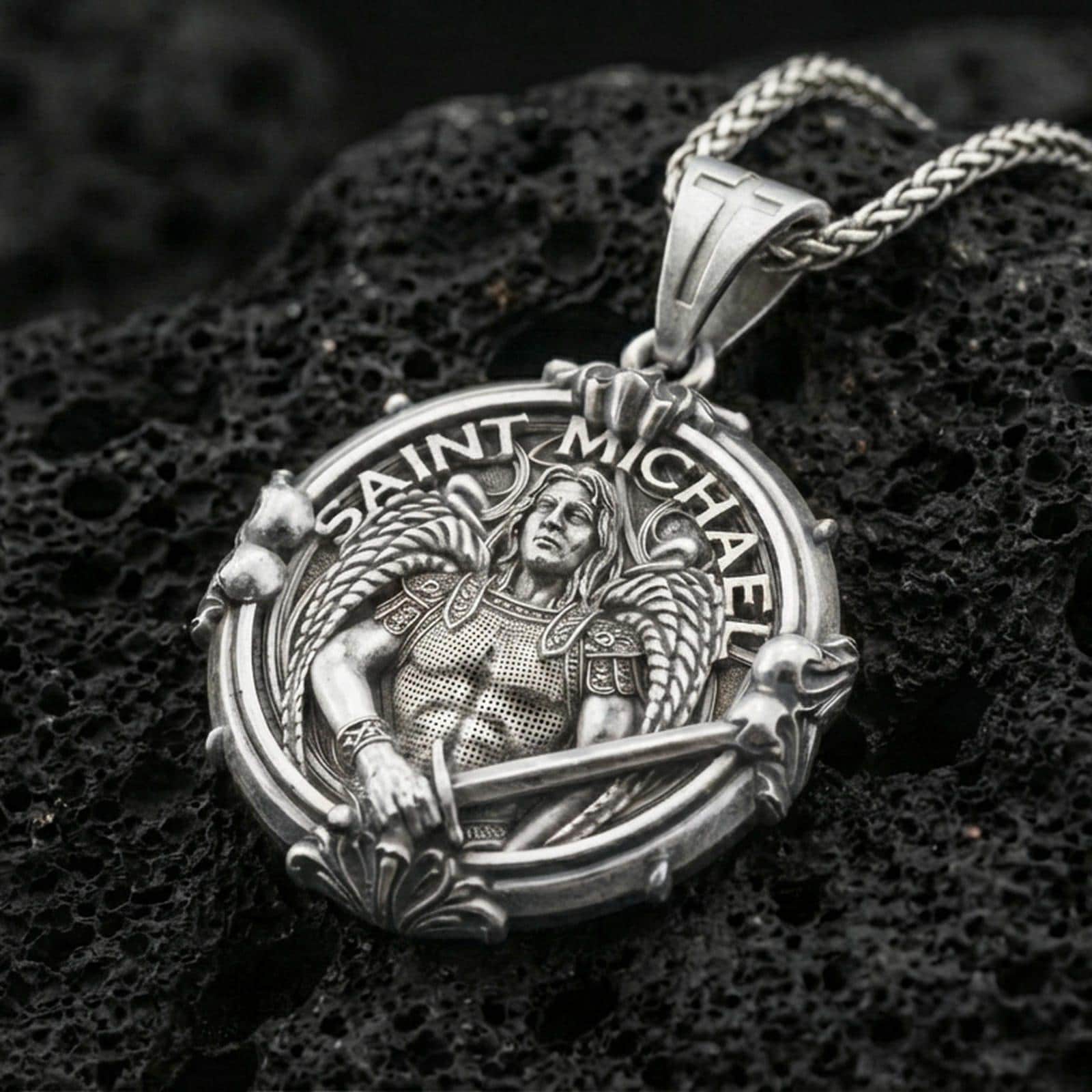Polished silver Archangel Michael medallion with round frame, armored angel relief, and decorative gothic border placed on textured volcanic rock in a premium studio product shot
