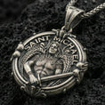 Load image into Gallery viewer, Oxidized Saint Michael pendant with winged warrior figure, sword detail, and Saint Michael inscription displayed on dark volcanic rock in a close-up luxury jewelry composition
