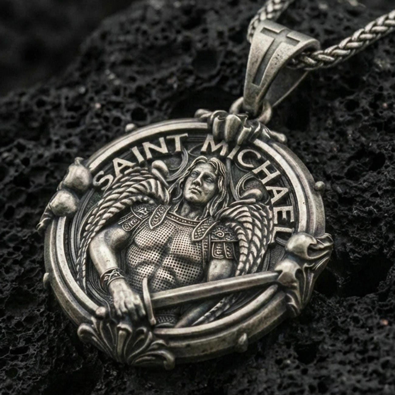 Oxidized Saint Michael pendant with winged warrior figure, sword detail, and Saint Michael inscription displayed on dark volcanic rock in a close-up luxury jewelry composition