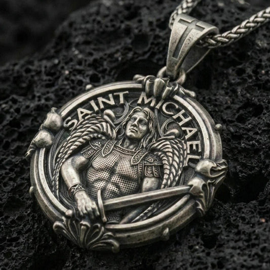 Oxidized Saint Michael pendant with winged warrior figure, sword detail, and Saint Michael inscription displayed on dark volcanic rock in a close-up luxury jewelry composition