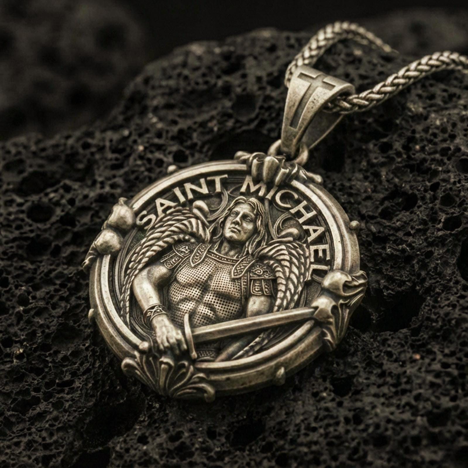 Bronze Saint Michael pendant with winged warrior figure, sword detail, and Saint Michael inscription displayed on dark volcanic rock in a close-up luxury jewelry composition