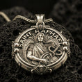 Load image into Gallery viewer, Bronze Saint Raphael pendant with winged archangel figure, healing staff with serpent, and Saint Raphael inscription displayed on dark volcanic rock in a close-up luxury jewelry composition
