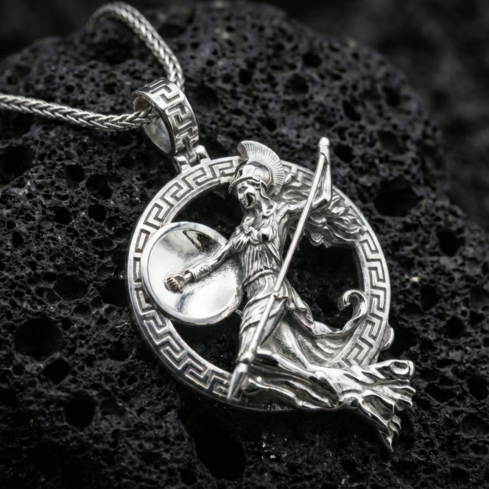 Polished silver Athena warrior pendant necklace on textured volcanic rock, crisp macro product photo emphasizing clean edges, sharp relief detailing on the shield and spear, and bright reflective highlights.