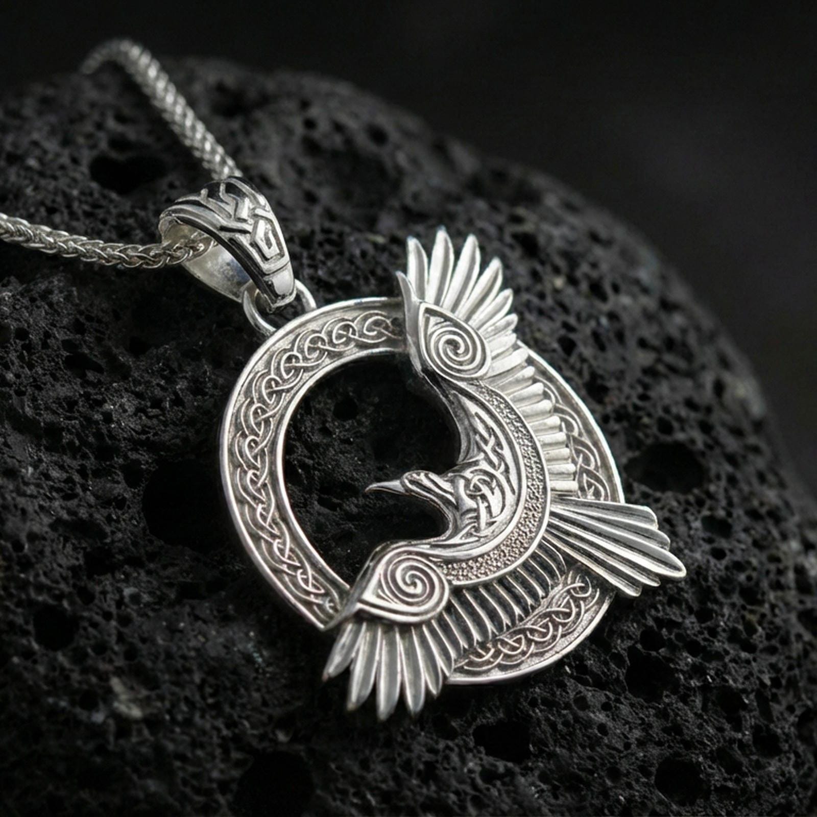 Polished silver Celtic raven knotwork pendant necklace on textured volcanic rock, crisp macro product photo emphasizing clean edges, sharp feather lines, and detailed Celtic weave engraving.