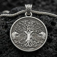 Load image into Gallery viewer, Oxidized Yggdrasil tree of life pendant necklace on textured volcanic rock, centered close-up highlighting the detailed branches, twin wolves, Celtic knot border, and deep antique shadowed contrast.
