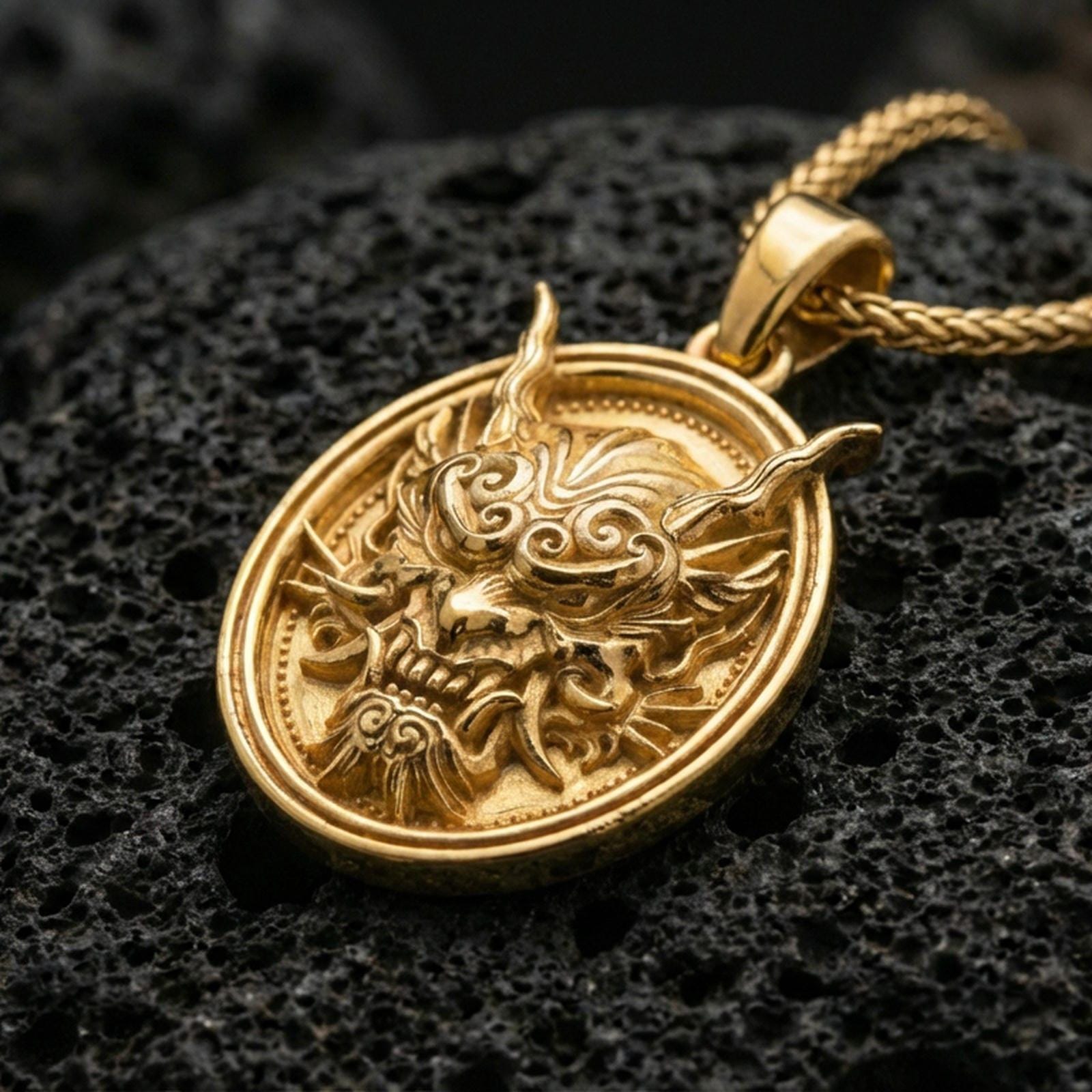 Gold plated demon mask medallion pendant necklace on textured volcanic rock, premium close-up with warm reflective shine across the horns, fierce expression, and raised circular frame.