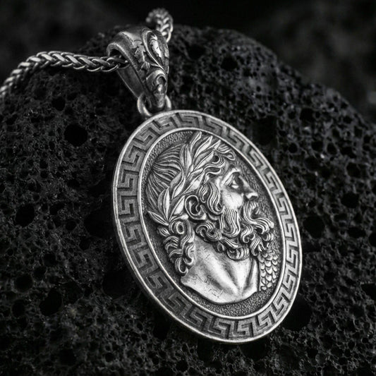 Oxidized Hades coin pendant necklace on textured volcanic rock, centered close-up highlighting the Greek key border, laurel-crowned profile, and deep antique shadowed contrast.