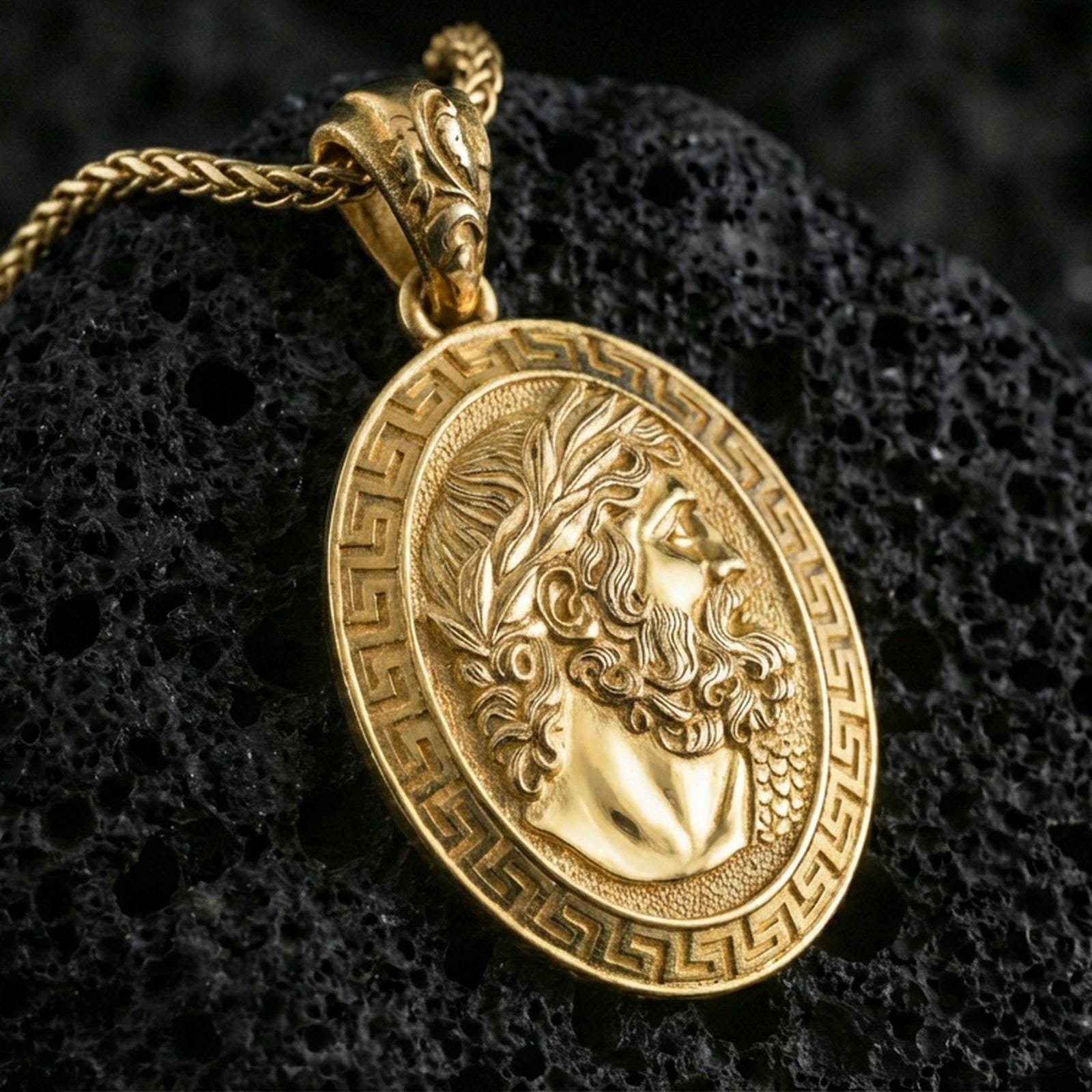 Gold plated Hades coin pendant necklace on textured volcanic rock, premium close-up with warm reflective shine across the laurel wreath, facial sculpting, and raised meander frame.