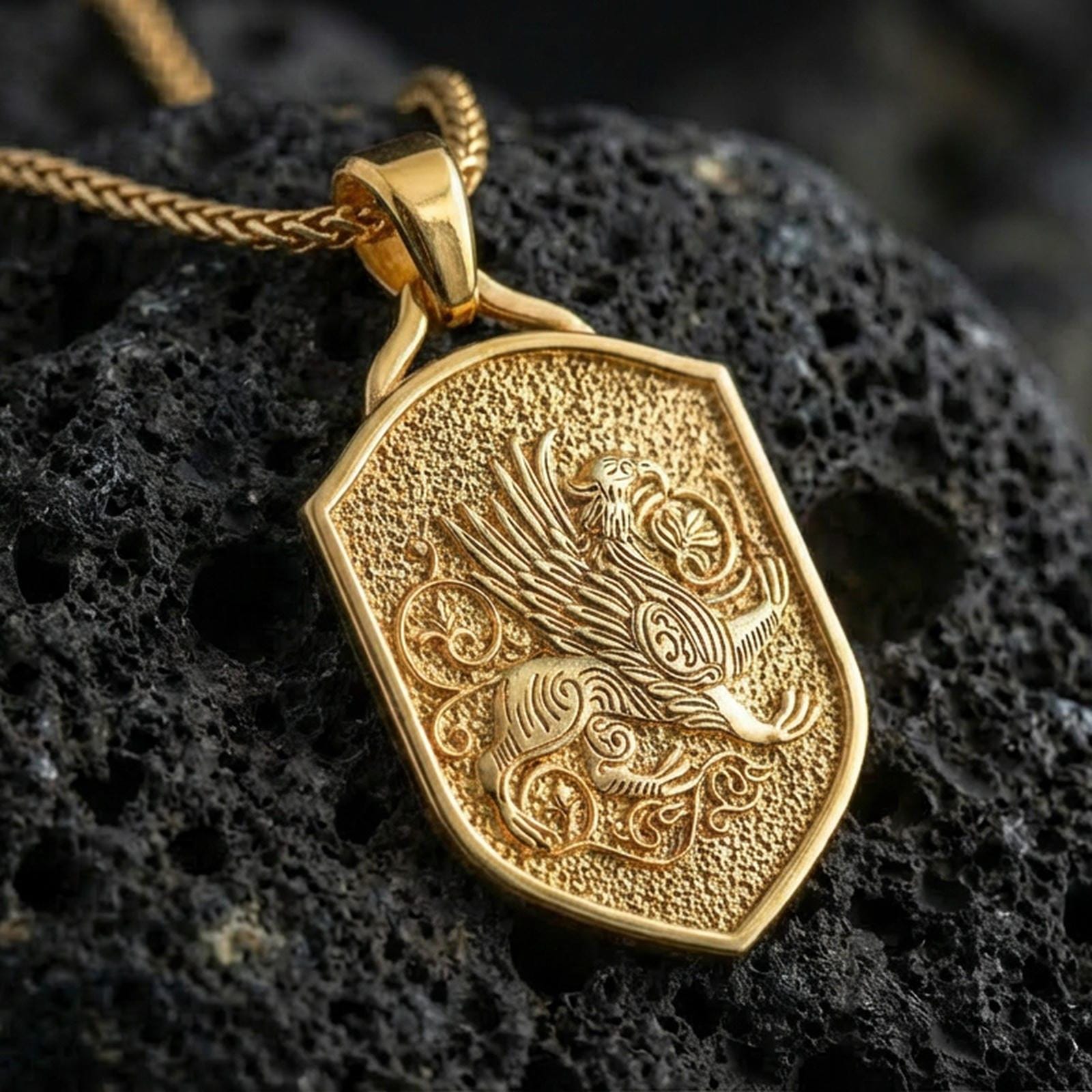 Gold plated griffin shield pendant necklace on textured volcanic rock, premium close-up with warm reflective shine across the wings, beak, and decorative scrollwork on the shield face.