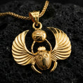 Load image into Gallery viewer, Gold plated winged scarab beetle pendant necklace on textured volcanic rock, premium close-up with warm reflective shine across the wings, scarab body, and raised Eye of Ra centerpiece.
