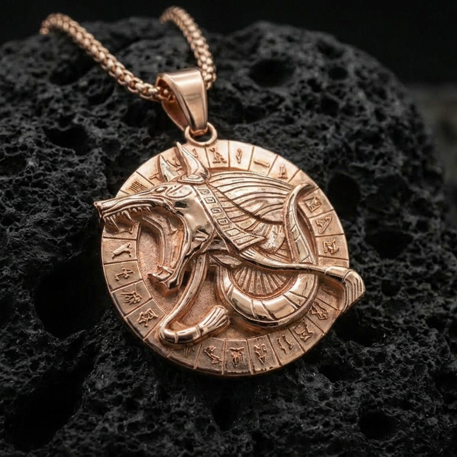 Rose gold plated Anubis medallion pendant necklace on textured volcanic rock, centered studio-style close-up showcasing soft rosy glow and intricate Egyptian jackal-and-hieroglyph detailing.