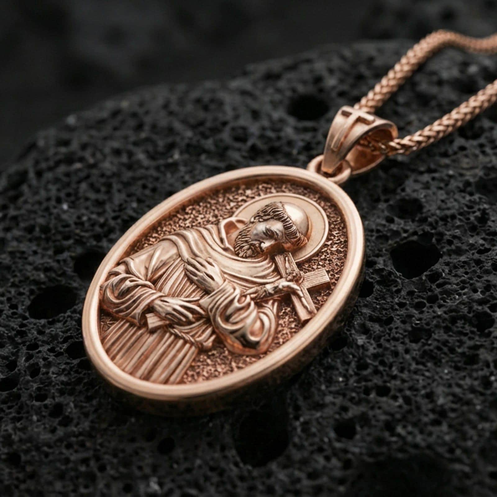 Rose gold plated oval Saint Francis necklace with sacred portrait relief, halo, and devotional cross resting on volcanic rock in an elegant close-up product presentation