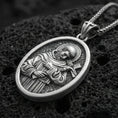 Load image into Gallery viewer, Oxidized Saint Francis pendant with halo, cross detail, and oval devotional portrait displayed on dark volcanic rock in a close-up luxury jewelry composition
