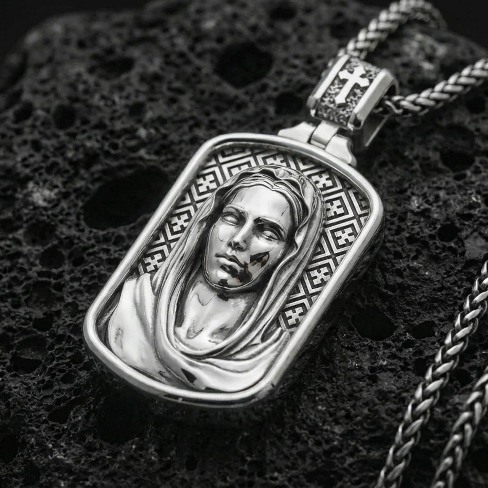 Polished silver Madonna medallion with detailed draped veil, calm facial relief, and framed devotional design placed on textured volcanic rock in a premium studio product shot