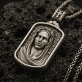 Load image into Gallery viewer, Bronze Virgin Mary pendant with serene veiled portrait and rounded rectangular frame displayed on dark volcanic rock in a close-up luxury jewelry composition
