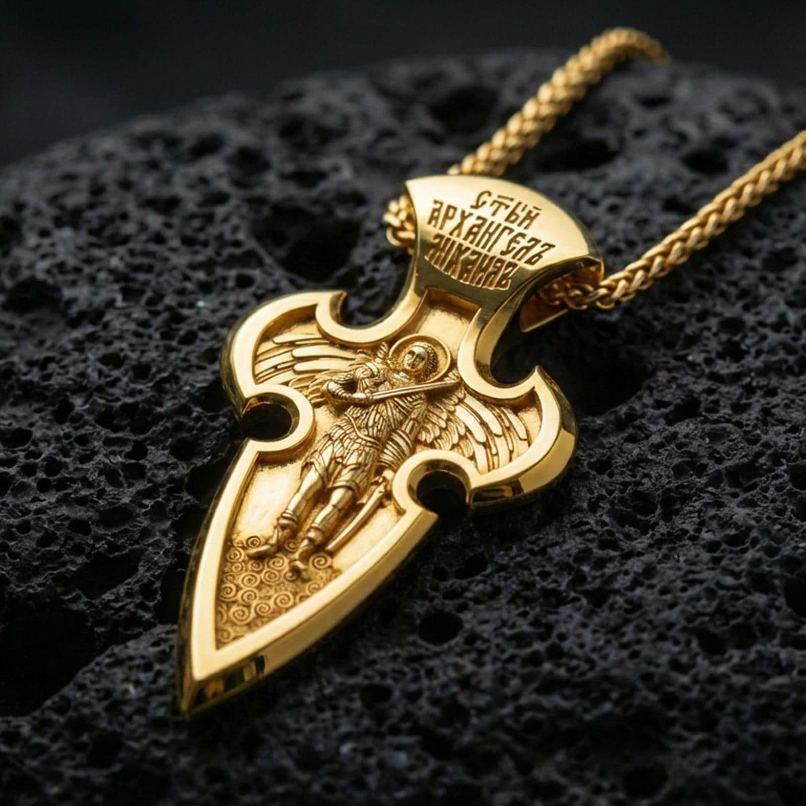 Gold plated Saint Michael pendant with winged protector motif, cross-shaped frame, and engraved inscription showcased on black volcanic rock in a refined macro jewelry image