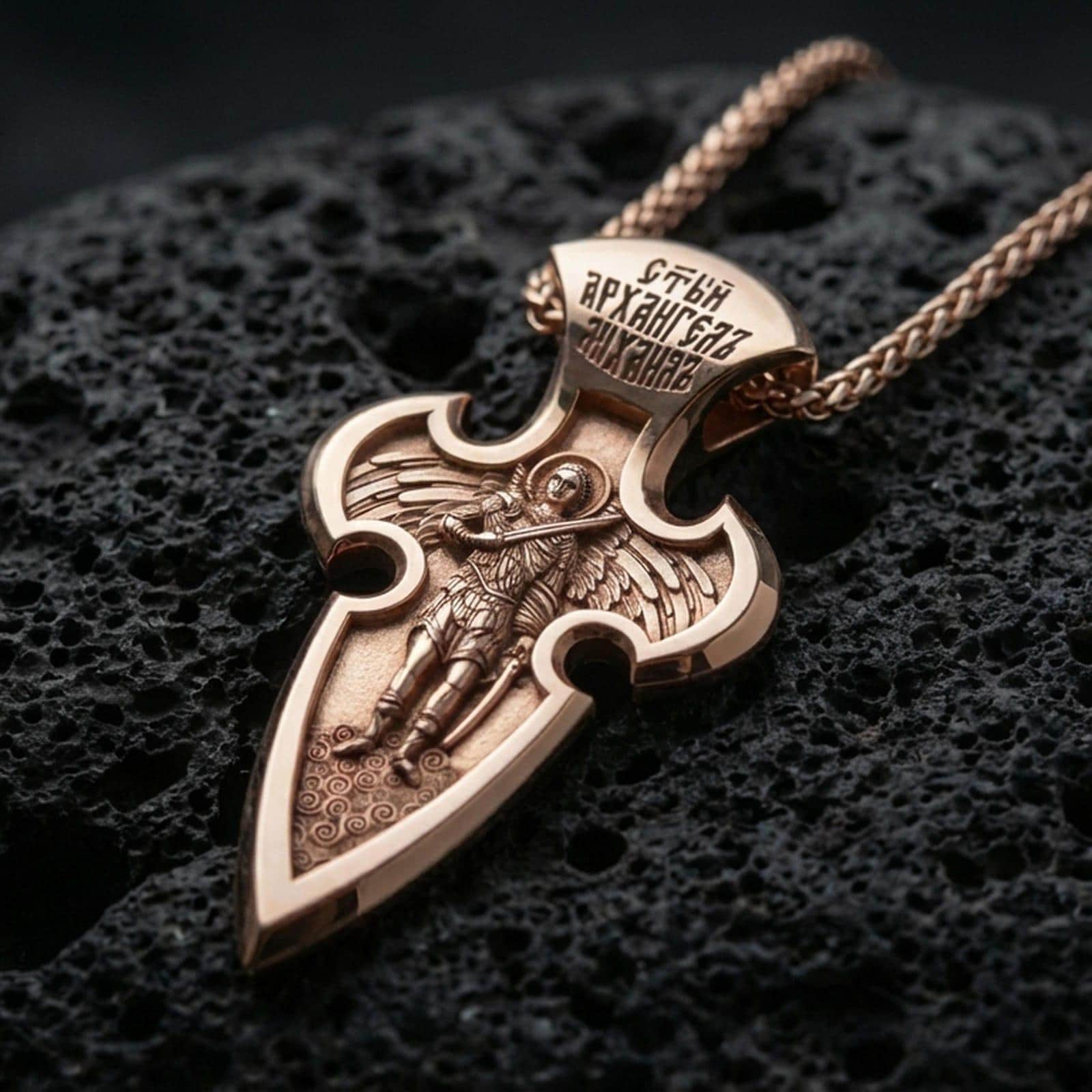 Rose gold plated Archangel Michael cross pendant with sacred warrior figure, elongated pointed silhouette, and detailed engraved top resting on volcanic rock in an elegant close-up product presentation