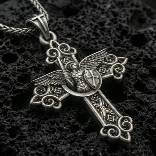 Oxidized Saint Michael cross pendant with winged archangel figure, shield detail, and ornate gothic scrollwork displayed on dark volcanic rock in a close-up luxury jewelry composition
