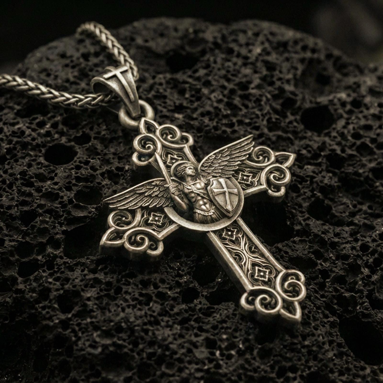 Bronze Saint Michael cross pendant with winged archangel figure, shield detail, and ornate gothic scrollwork displayed on dark volcanic rock in a close-up luxury jewelry composition