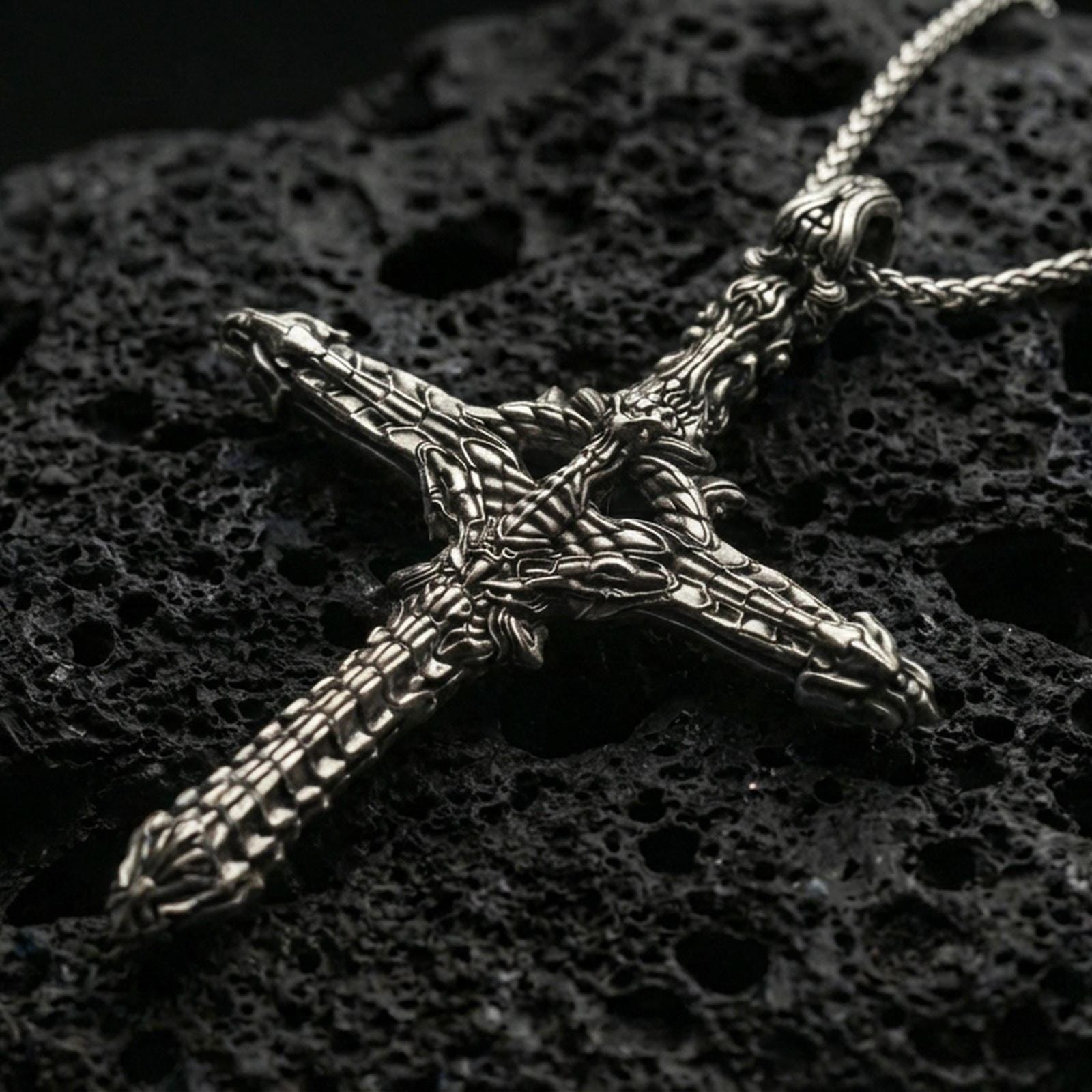 Oxidized biomechanical gothic cross pendant with intricate skeletal and machine-like detailing displayed on dark volcanic rock in a close-up luxury jewelry composition