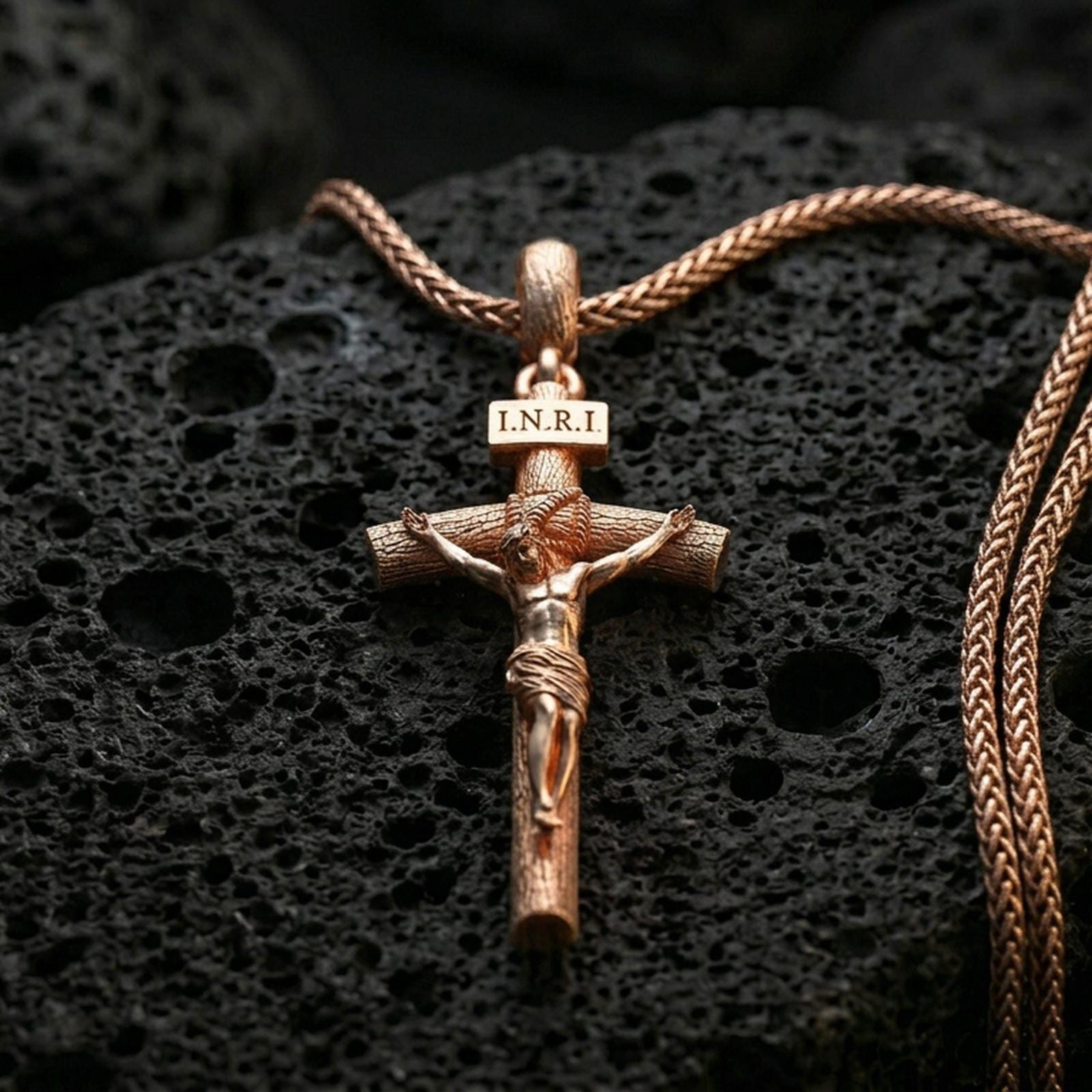 Rose gold plated Jesus cross pendant with sculpted corpus, INRI inscription, and slender crucifix form resting on volcanic rock in an elegant close-up product presentation
