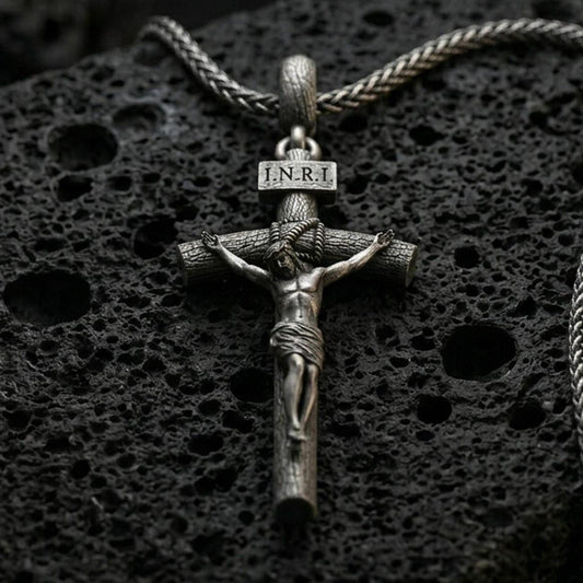 Oxidized INRI crucifix pendant with sculpted Jesus figure and wood-textured cross displayed on dark volcanic rock in a close-up luxury jewelry composition